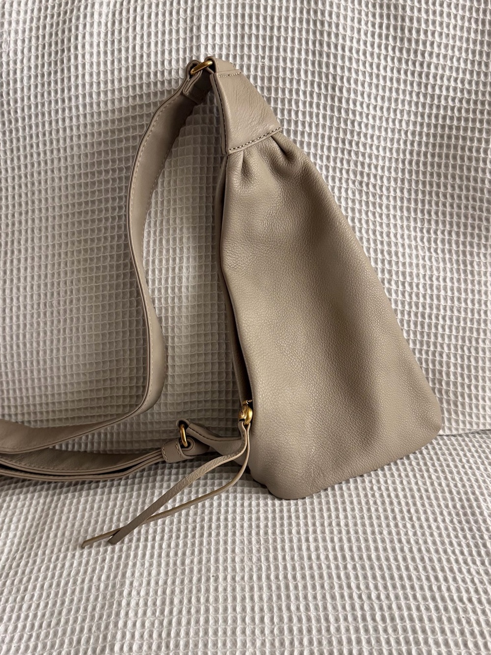 taupe leather sling bag by HOBO next to its matching cream-colored dust bag